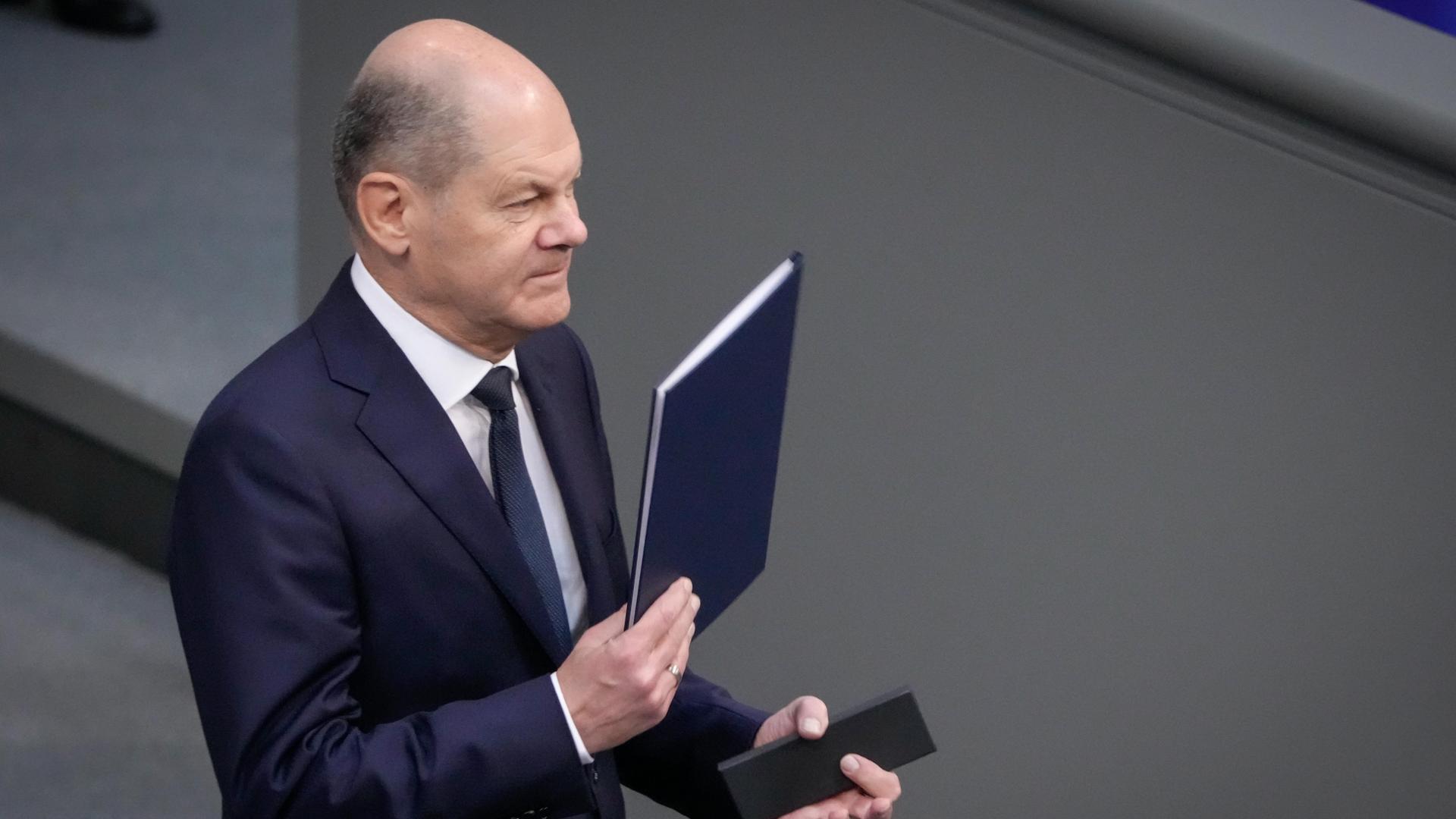 Bundeskanzler Olaf Scholz hält im Bundestag eine Mappe mit Blättern in der Hand.