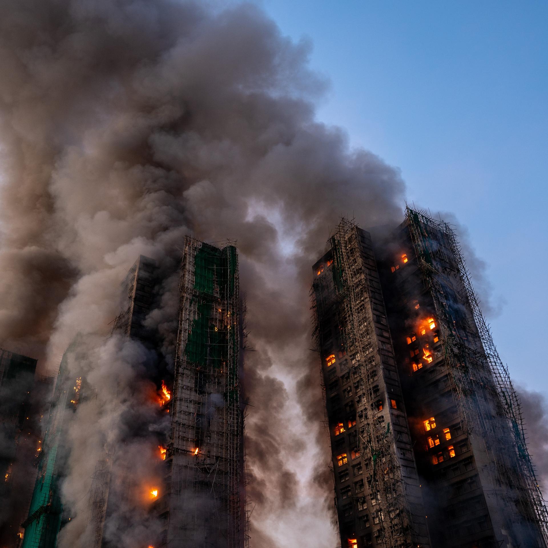 Die drei nebeneinander stehenden Hochhauser sind in dichten schwarzen Rauch gehüllt. Aus vielen Fenster lodern Flammen. Darüber ein dunkelblauer Himmel.