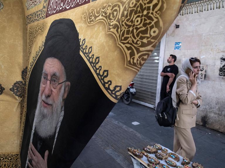 Alltag im Iran. Eine Frau geht in Teheran an einer Flagge mit dem Porträt des bei Luftangriffen der USA und Israels getöteten geistlichen Führers Ayatollah Ali Chamenei vorbei