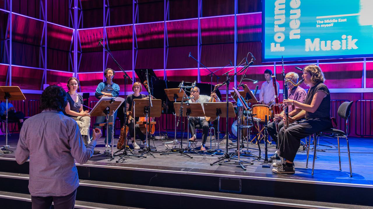 Das Ensemble Aventure sitzt auf einer blau-lila beleuchteten Bühne. Ihre Konzentration ist auf einen Mann gerichtet, der vor der erhöhten Bühne steht und mit den Musikerinnen und Musikern interagiert.