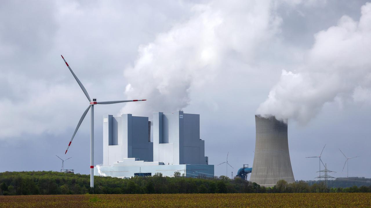 Hinter einer grünen Wiese steht das RWE-Kraftwerk in Grevenbroich mit Kühltürmen aus denen Rauch in den Himmel steigt. Davor und dahinter stehen Windräder.