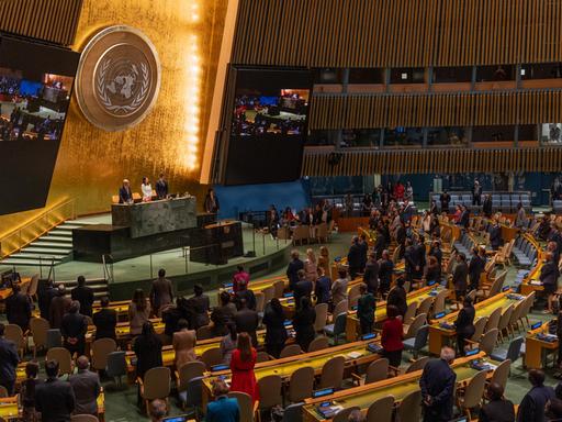 Blick in den Plenarsaal der UNO in New York, wo die Delegierten stehen. An der goldfarbenen Rückwand sieht man das Emblem der Organisation.