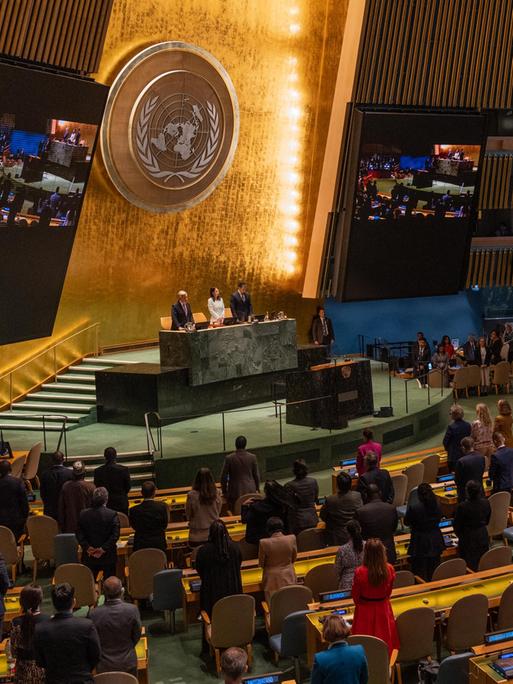 Blick in den Plenarsaal der UNO in New York, wo die Delegierten stehen. An der goldfarbenen Rückwand sieht man das Emblem der Organisation.