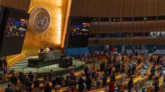 Blick in den Plenarsaal der UNO in New York, wo die Delegierten stehen. An der goldfarbenen Rückwand sieht man das Emblem der Organisation.