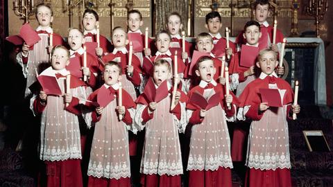 Jungs halten Kerzen und Notenblätter in den Händen und singen in einer Kirche.