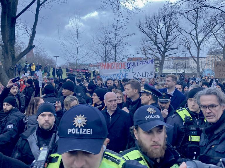Demo im Görlitzer Park: Berlins Regierdender Bürgermeister Kai Wegner steht in einer Menge zwischen Polizei und Demonstrierenden. Im Hintergrund ein Banner mit der Aufschrift "Feministische Schutzräume statt populistische Zäune".