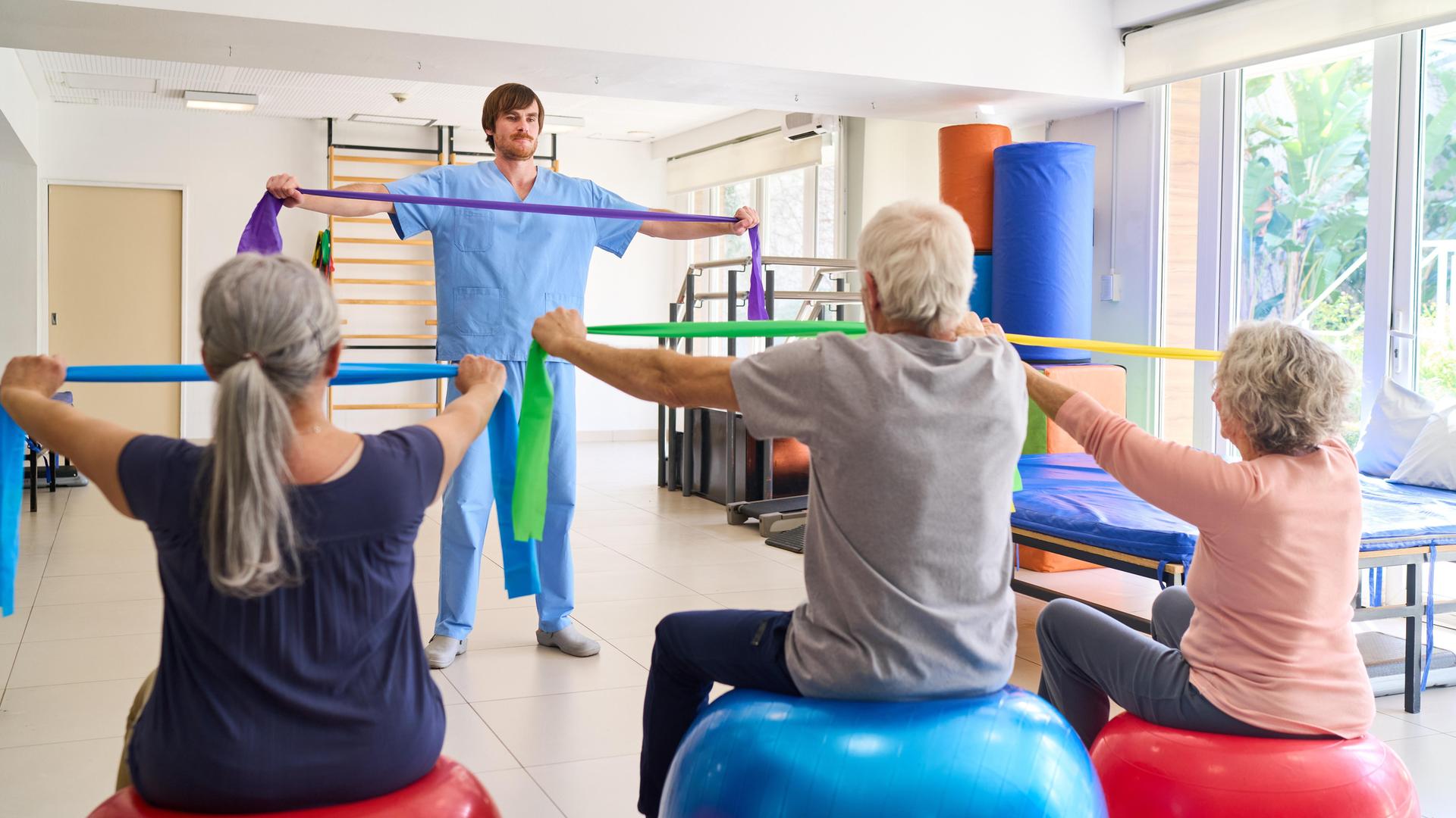 Zwei ältere Frauen und ein älterer Mann üben in einem Gymnastikkurs mit elastischen Bändern. Vor ihnen steht ein jüngerer Mann und zeigt die Übung.