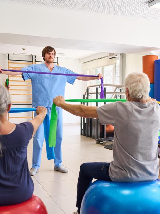 Zwei ältere Frauen und ein älterer Mann üben in einem Gymnastikkurs mit elastischen Bändern. Vor ihnen steht ein jüngerer Mann und zeigt die Übung.
