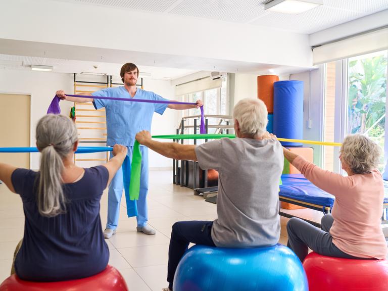 Zwei ältere Frauen und ein älterer Mann üben in einem Gymnastikkurs mit elastischen Bändern. Vor ihnen steht ein jüngerer Mann und zeigt die Übung.