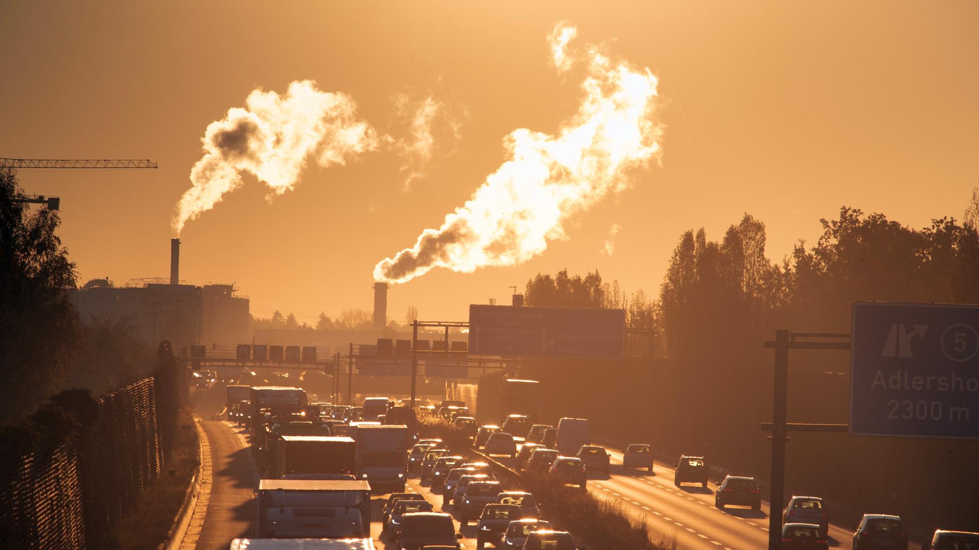 Stau am Morgen auf der Autobahn, im Gegenlicht sind im Hintergrund Rauschschwaden aus Industrieschornsteinen zu erkennen.