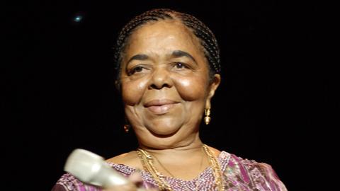 2006 steht Cesária Évora in einer lilafarbenen Bluse mit Mikrophon in den Händen auf einer Bühne und lächelt.