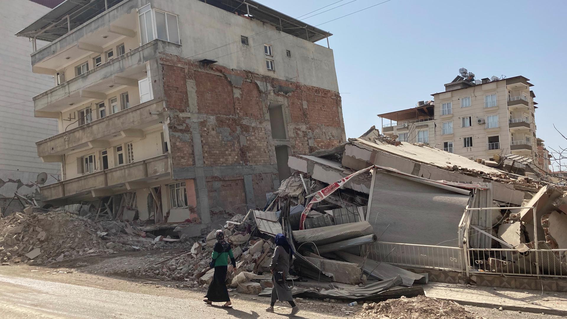 Zwei Frauen gehen auf einer Straße an zerstörten Häusern im türkischen Ort Kirikhan nahe der syrischen Grenze vorbei, knapp einen Monat nach den schweren Erdbeben.