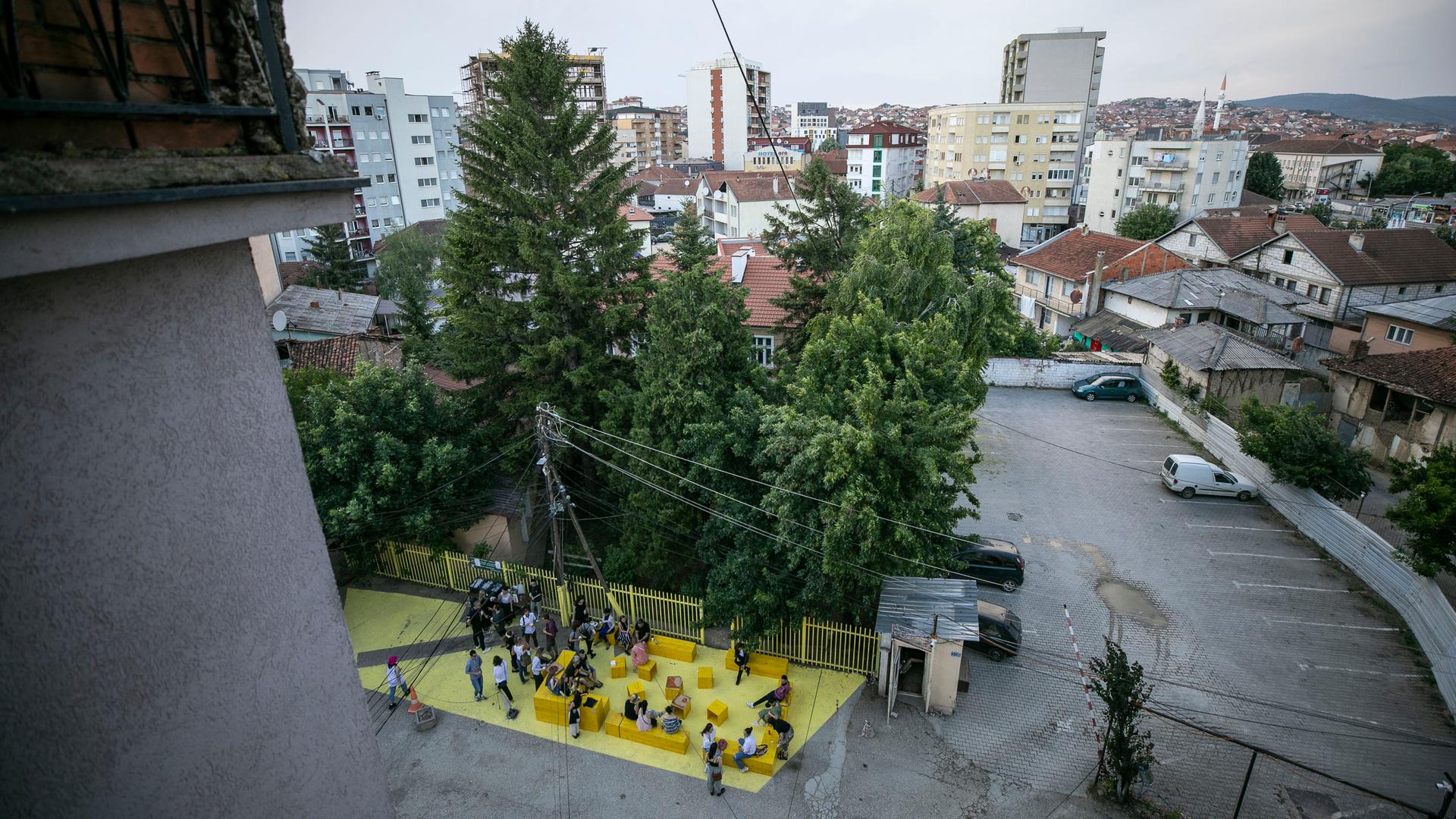 Blick über Pristina und einen Innenhof, wo gelbe Sitzblöcke stehen, auf denen Menschen sitzen. 