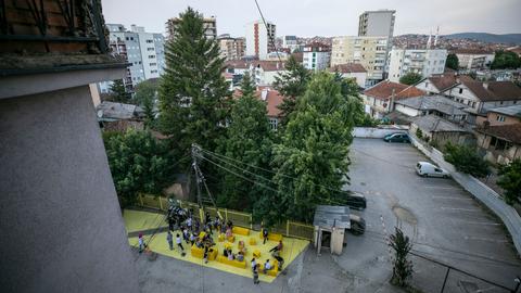 Blick über Pristina und einen Innenhof, wo gelbe Sitzblöcke stehen, auf denen Menschen sitzen. 