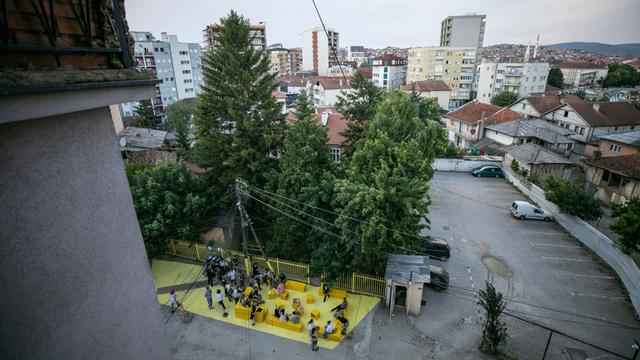 Blick über Pristina und einen Innenhof, wo gelbe Sitzblöcke stehen, auf denen Menschen sitzen. 