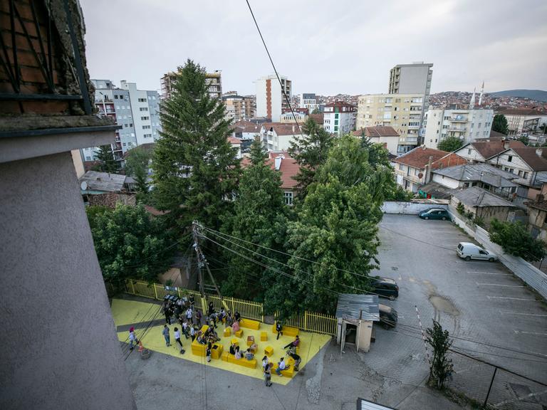 Blick über Pristina und einen Innenhof, wo gelbe Sitzblöcke stehen, auf denen Menschen sitzen. 