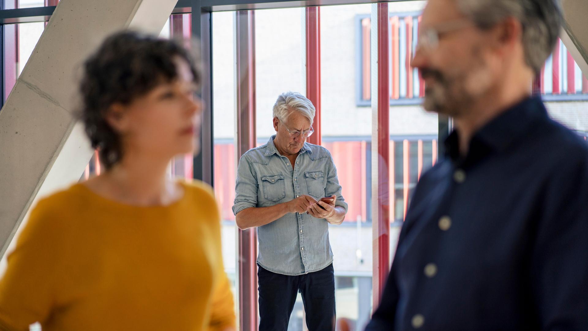 Eine Frau und ein Mann reden im Vordergrund, im Hintergrund steht ein Mann mit einem Smartphone