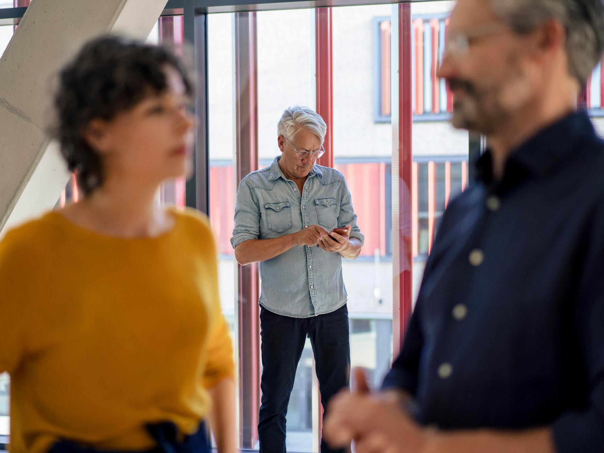 Eine Frau und ein Mann reden im Vordergrund, im Hintergrund steht ein Mann mit einem Smartphone