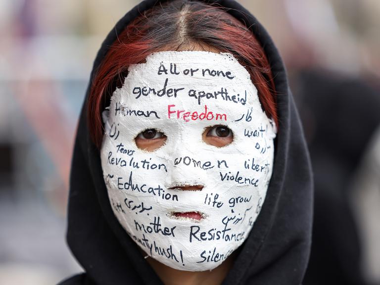 Eine Frau trägt eine Gipsmaske, auf der unter anderem steht: "All or none. Gender Arpartheid. Freedom. Eduaction. Resistance" 