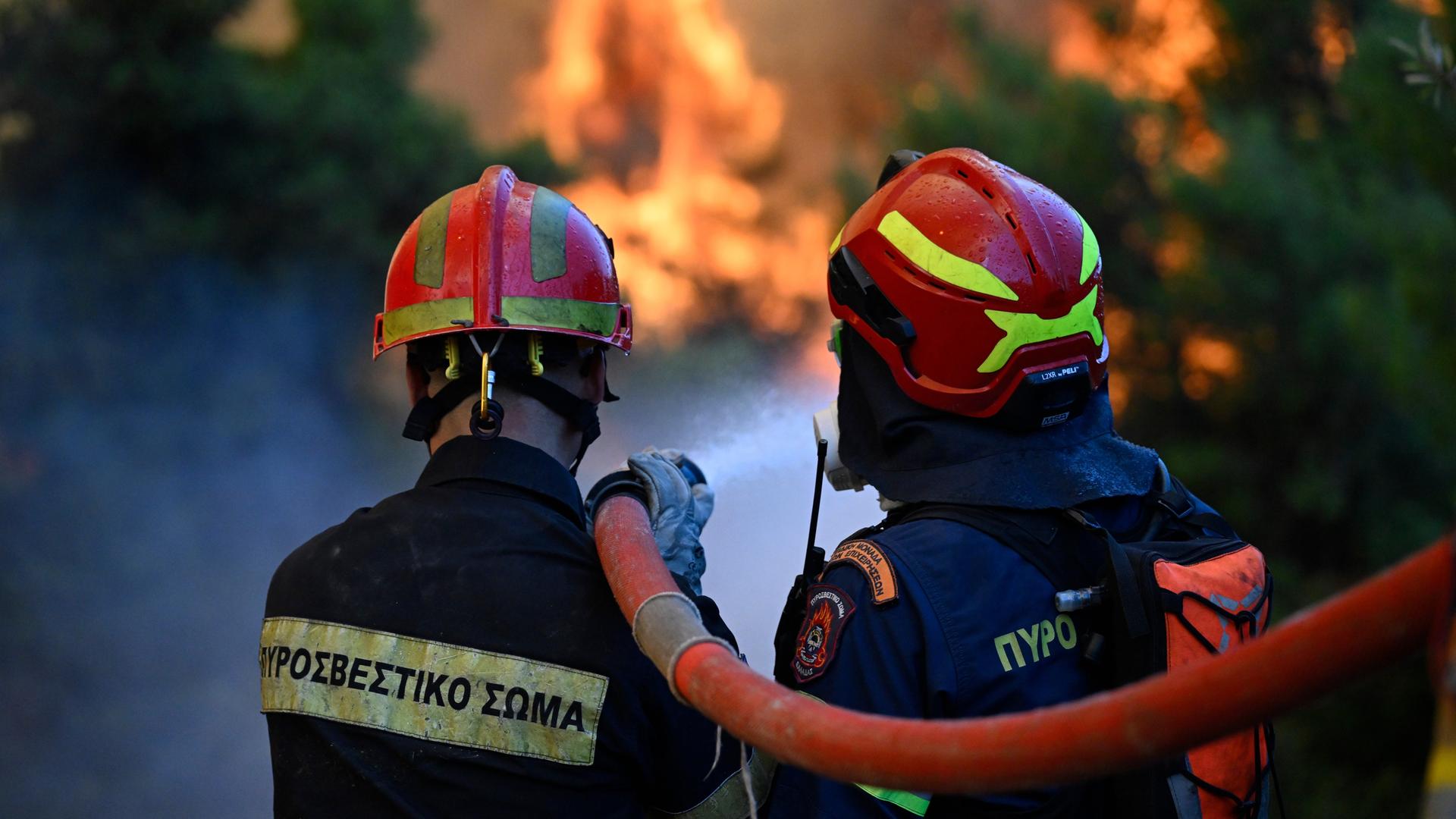Zwei Feuerwehrleute stehen nebeneinander und halten einen Wasserschlauch. Sie blicken auf die Flammen. Zwei Feuerwehrleute stehen nebeneinander und halten einen Wasserschlauch. Sie blicken auf die Flammen.