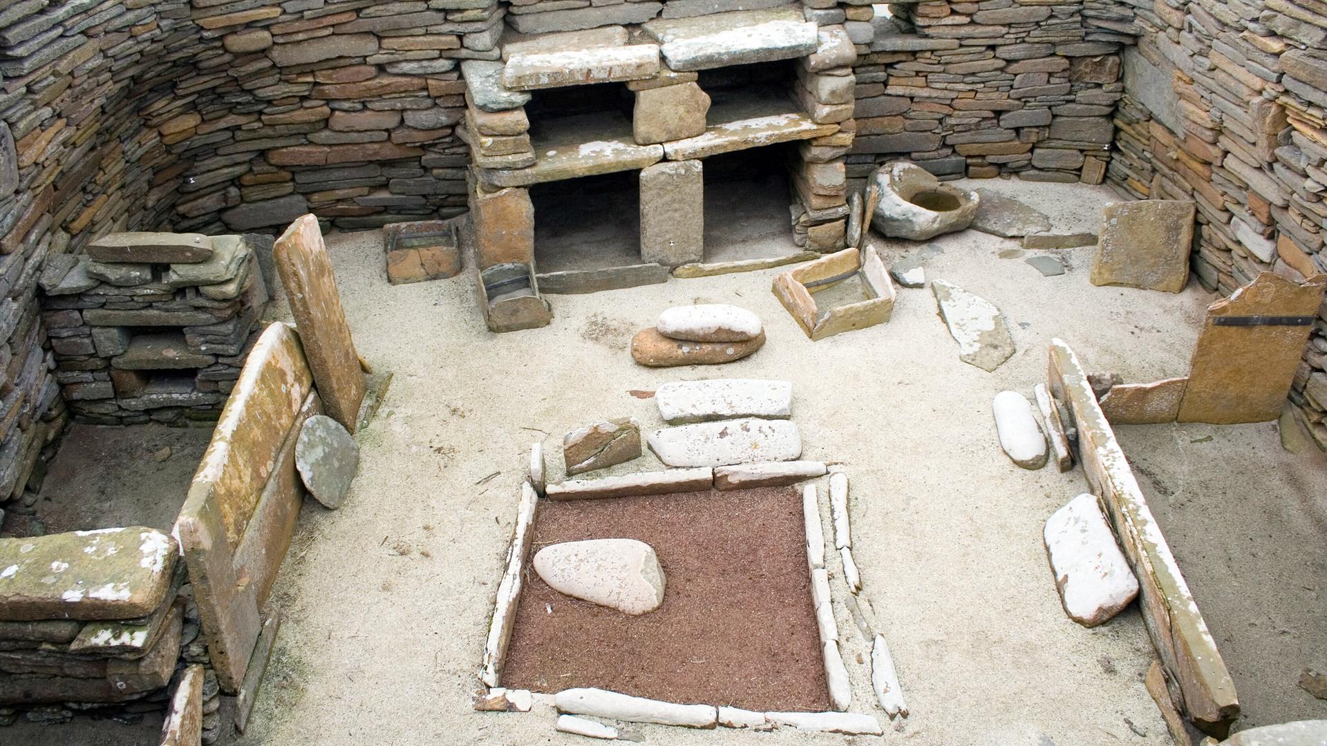 Ein jungsteinzeitliches Haus in der Ausgrabungsstätte Skara Brae in Schottland. Das Gebäude aus dem Neolithikum zeigt links und rechts Steinkistenbetten, in der Mitte eine Feuerstelle und hinten ein Regal. Ein jungsteinzeitliches Haus in der Ausgrabungsstätte Skara Brae in Schottland. Das Gebäude aus dem Neolithikum zeigt links und rechts Steinkistenbetten, in der Mitte eine Feuerstelle und hinten ein Regal.
