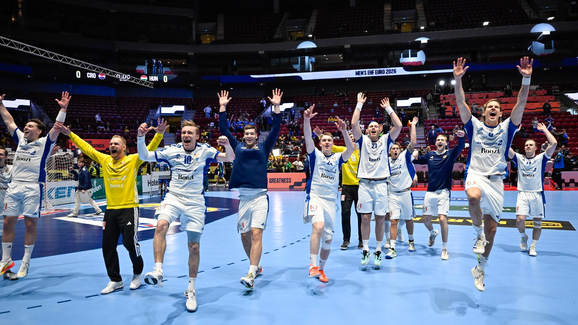 Die Spieler in weißen Trikots hüpfen auf dem blauen Feld mit erhobenen Armen. Dahinter Zuschauertribünen.