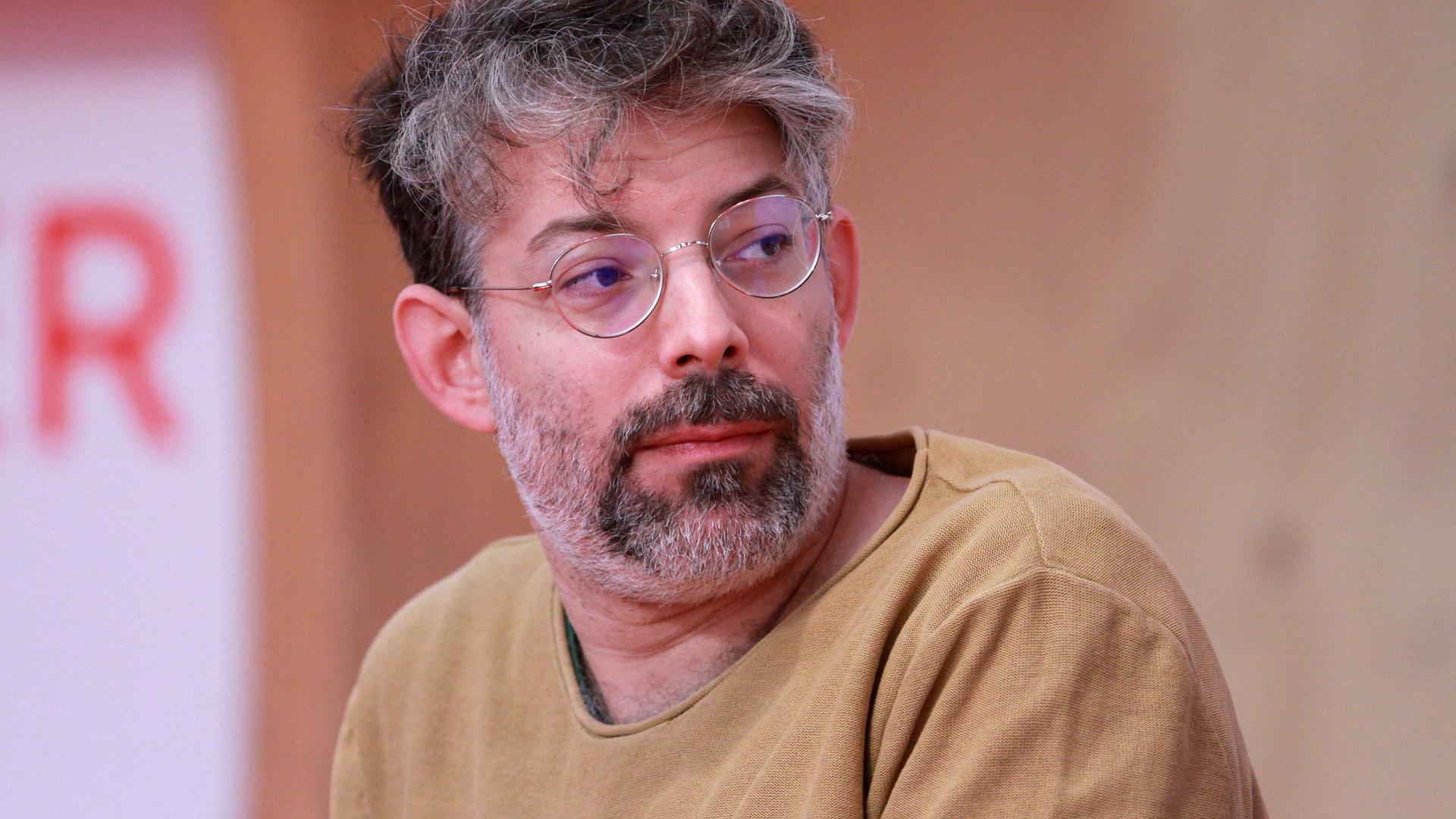 Der Autor Tomer Dotan-Dreyfus sitzt auf einem Podium, trägt einen sandfarbenen Pullover und schaut zur Seite.