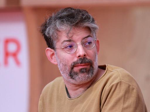 Der Autor Tomer Dotan-Dreyfus sitzt auf einem Podium, trägt einen sandfarbenen Pullover und schaut zur Seite.