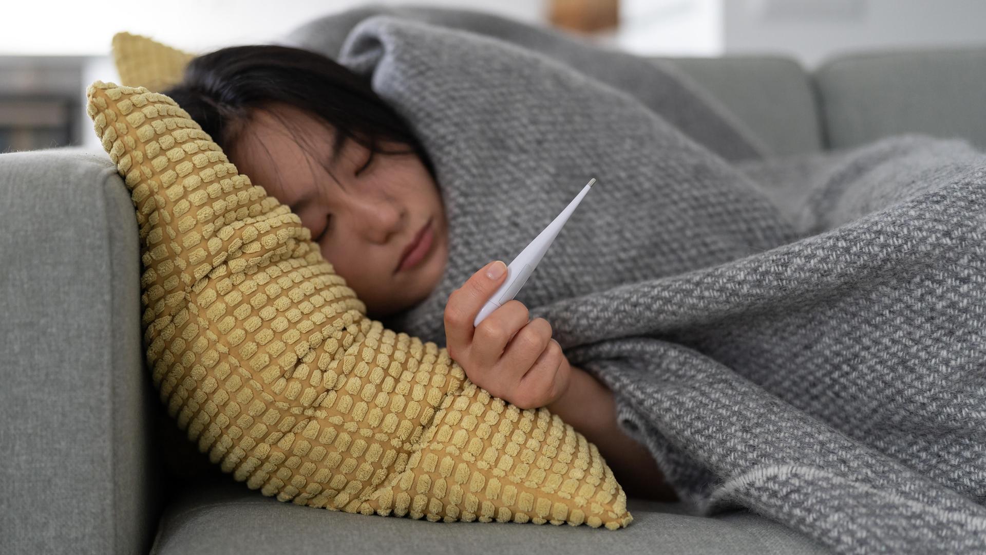 Eine Frau liegt eingemümmelt auf dem Sofa, sie hält ein Fieberthermometer in der Hand, sie ist krank