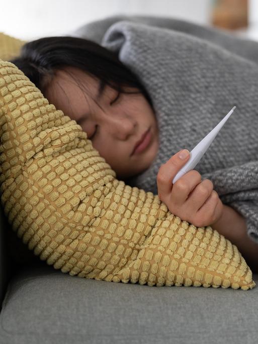 Eine Frau liegt eingemümmelt auf dem Sofa, sie hält ein Fieberthermometer in der Hand, sie ist krank