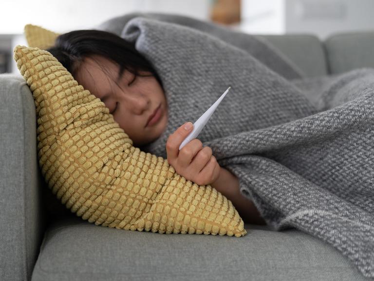 Eine Frau liegt eingemümmelt auf dem Sofa, sie hält ein Fieberthermometer in der Hand, sie ist krank
