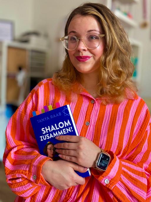 Tanya Yael Raab trägt eine rosaorange farbene Bluse und hält ihr Buch "Shalom zusammen!" im Arm.
