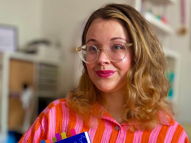 Tanya Yael Raab trägt eine rosaorange farbene Bluse und hält ihr Buch "Shalom zusammen!" im Arm.