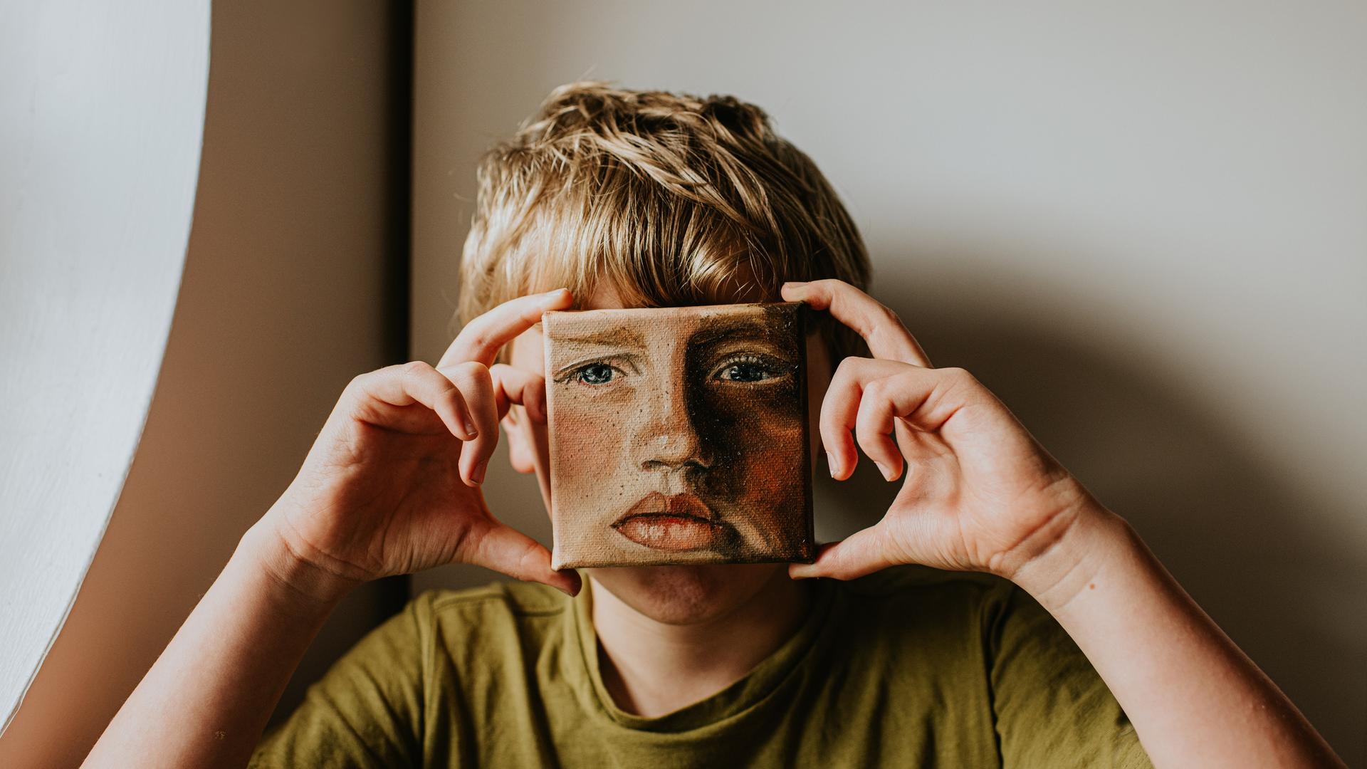 Ein Junge bedeckt sein Gesicht mit einem kleinen Ölgemälde von sich selbst, das Gesicht im Bild hat einen ernsten Ausdruck Ein Junge bedeckt sein Gesicht mit einem kleinen Ölgemälde von sich selbst, das Gesicht im Bild hat einen ernsten Ausdruck