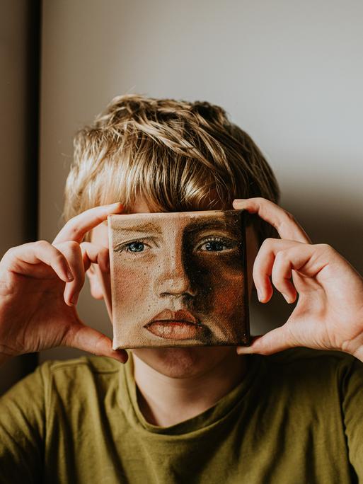 Ein Junge bedeckt sein Gesicht mit einem kleinen Ölgemälde von sich selbst, das Gesicht im Bild hat einen ernsten Ausdruck