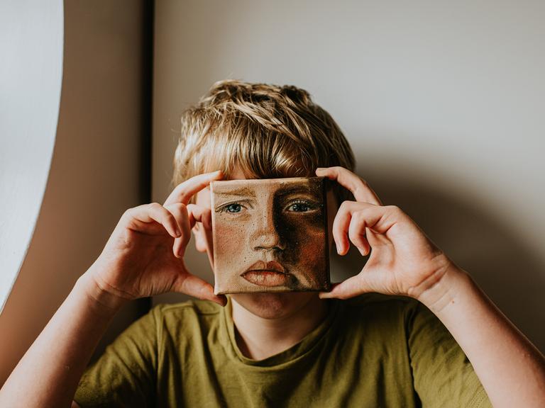 Ein Junge bedeckt sein Gesicht mit einem kleinen Ölgemälde von sich selbst, das Gesicht im Bild hat einen ernsten Ausdruck