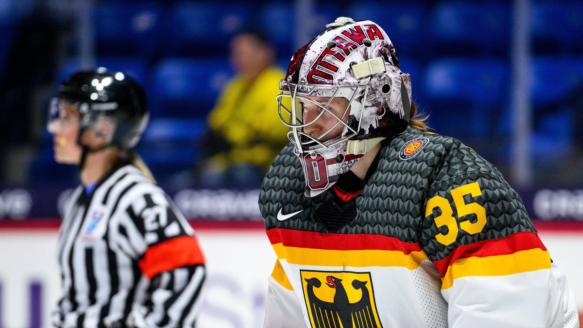 Torhüterin Sandra Abstreiter bei einem Eishockey-Spiel mit Helm und weißem National-Trikot mit Bundesadler. Im Hintergrund sieht man eine Schiedsrichterin. Torhüterin Sandra Abstreiter bei einem Eishockey-Spiel mit Helm und weißem National-Trikot mit Bundesadler. Im Hintergrund sieht man eine Schiedsrichterin.