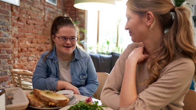 Eine Frau mit Down-Syndrom mit ihrer Freundin in einem Cafe beim Gespräch. Vor ihnen stehen belegte Toasts. 