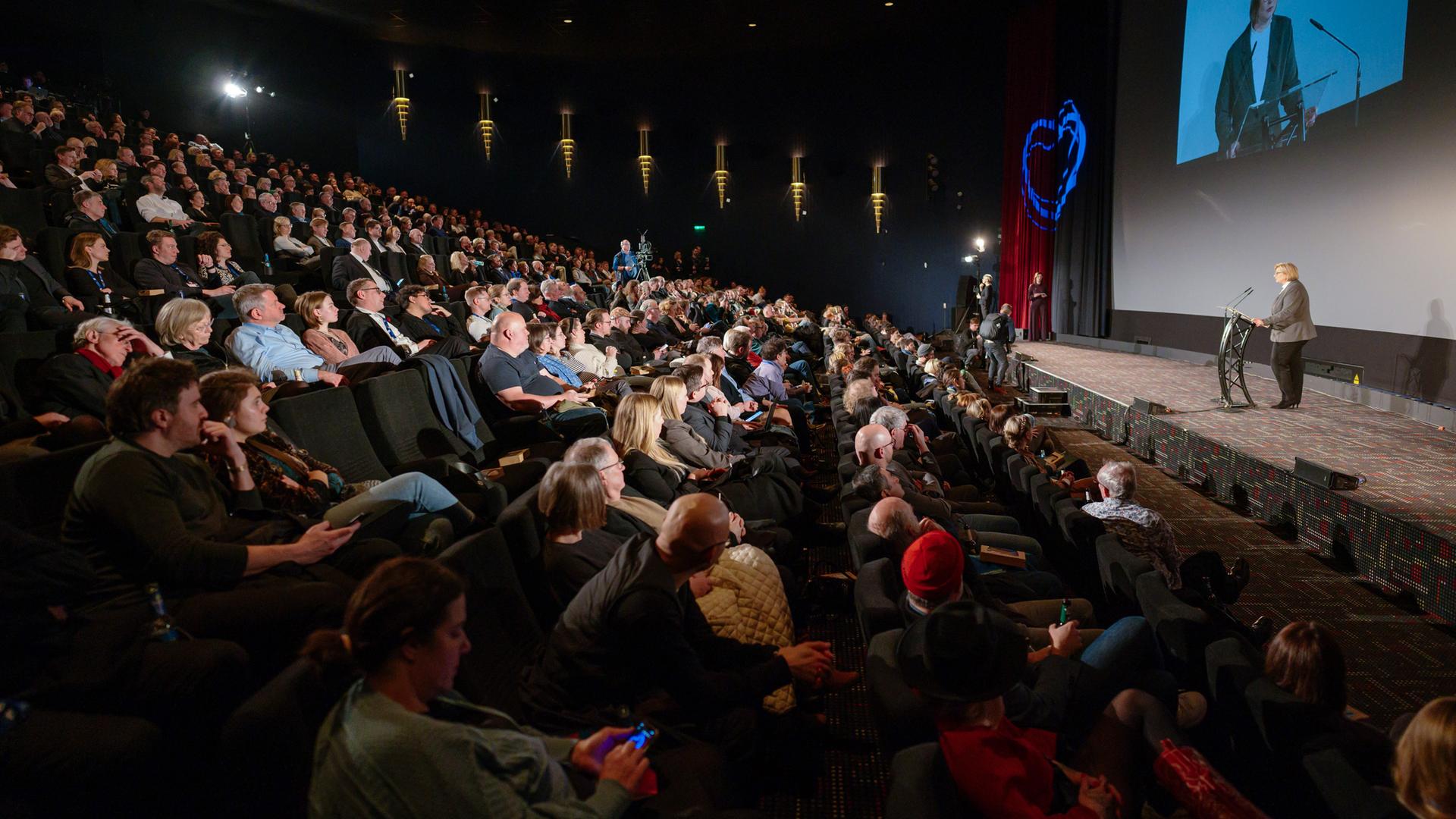 Blick in einen vollbesetzten Kinosaal. Auf der Bühne hält ein Mann eine Rede.