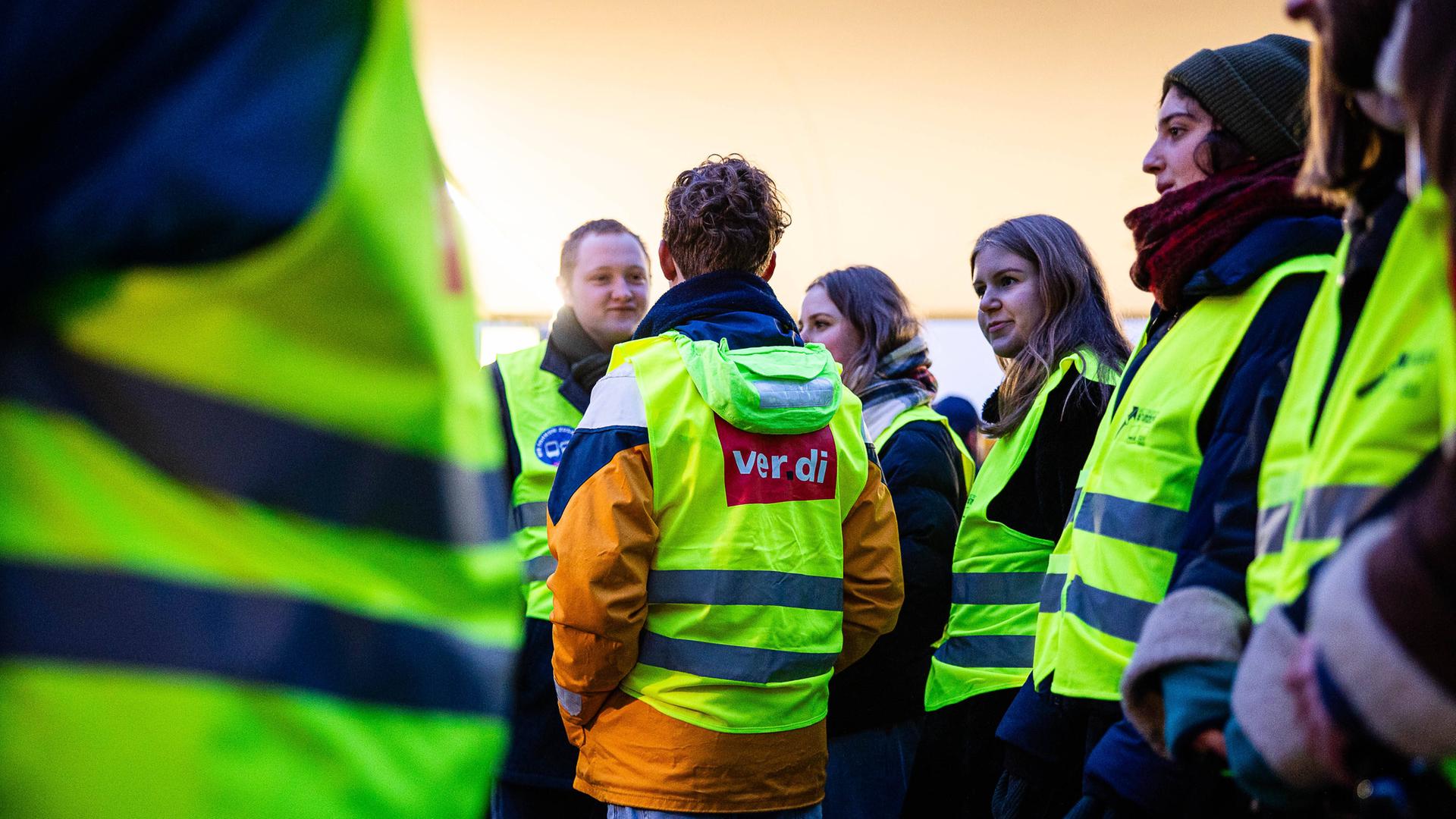 Menschen in gelb-leuchtenden Warn-Westen mit Ver-Di-Aufdruck stehen zusammen. Menschen in gelb-leuchtenden Warn-Westen mit Ver-Di-Aufdruck stehen zusammen.