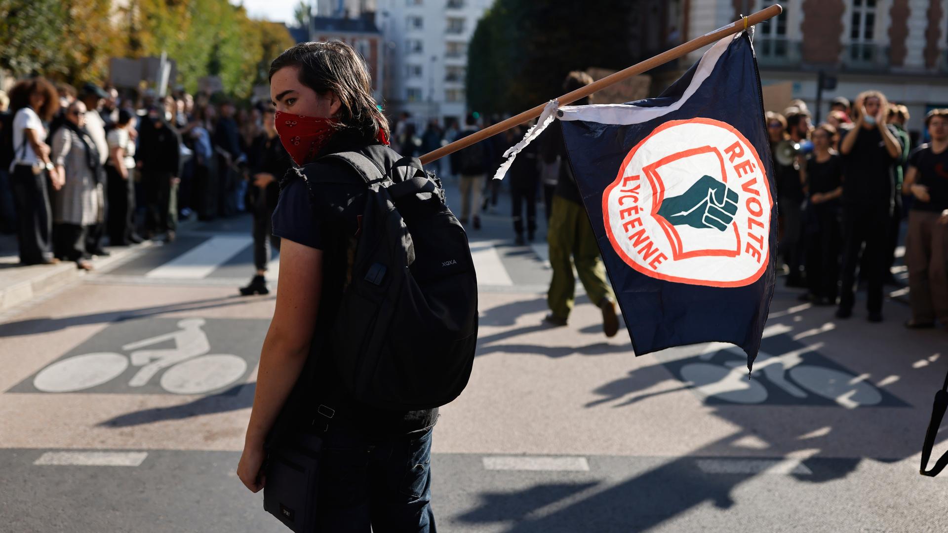 Eine vermummte junge Person trägt eine Fahne am Rand einer großen Demonstration. Eine vermummte junge Person trägt eine Fahne am Rand einer großen Demonstration.