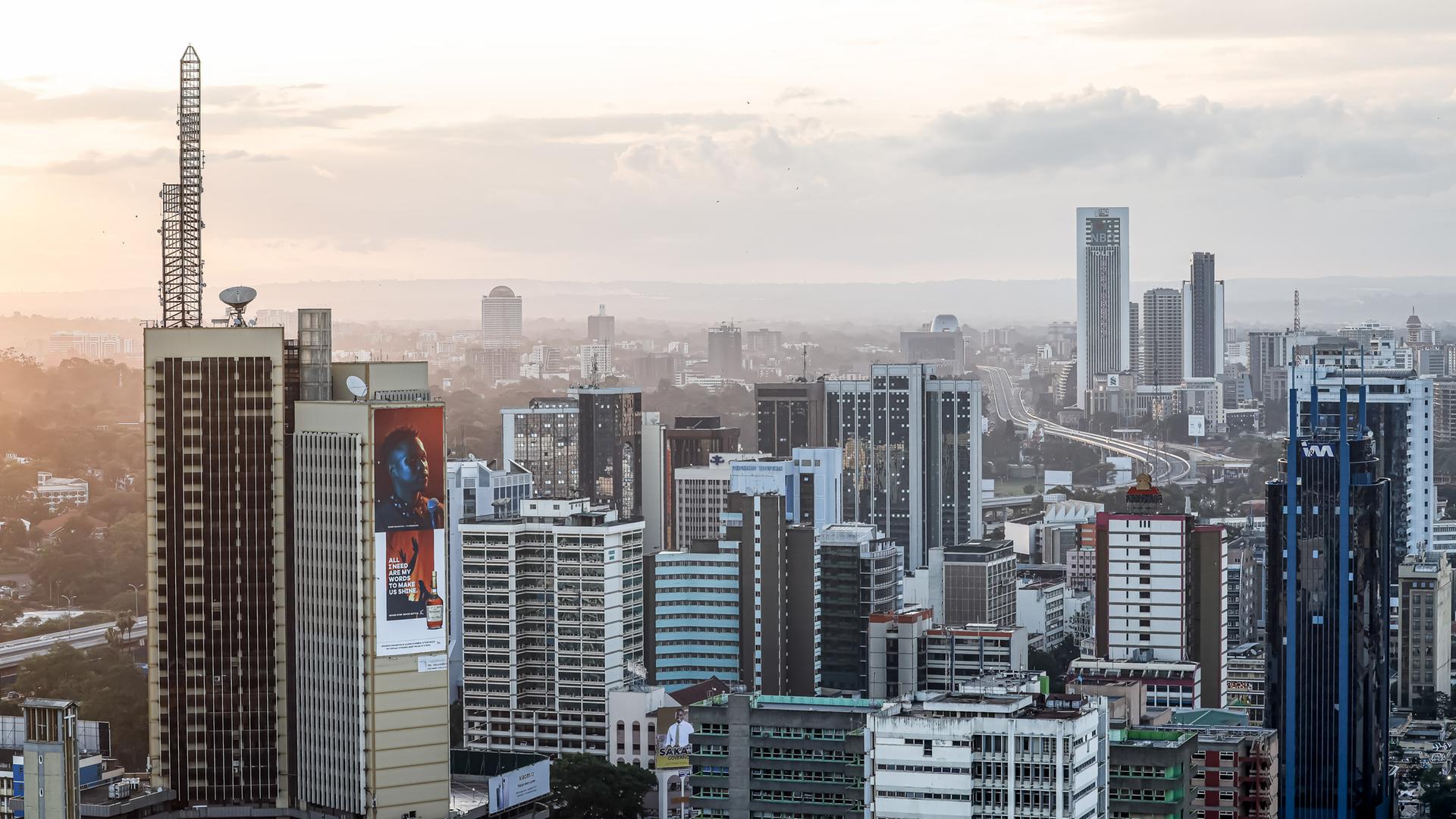 Zu sehen ist eine Luftaufnahme von Nairobi. Im Vorder- sowie im Hintergrund stehen mehrere Hochhäuser. Dazwischen schlängelt sich eine Straße. Zu sehen ist eine Luftaufnahme von Nairobi. Im Vorder- sowie im Hintergrund stehen mehrere Hochhäuser. Dazwischen schlängelt sich eine Straße.