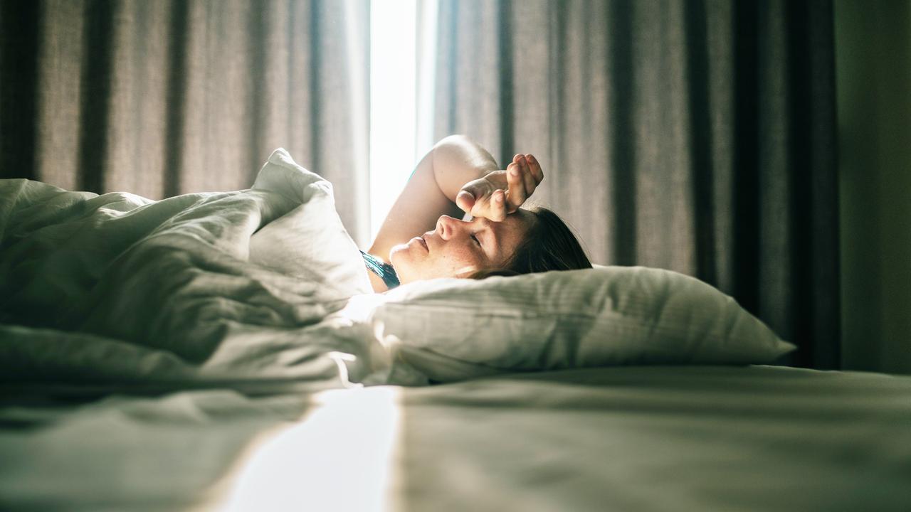 Eine Frau liegt im Bett. Sonniges Tageslicht fällt durch die Vorhänge im Hintergrund. Sie hatte eine Hand an die Stirn gelegt.