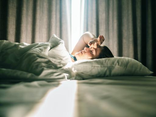 Eine Frau liegt im Bett. Sonniges Tageslicht fällt durch die Vorhänge im Hintergrund. Sie hatte eine Hand an die Stirn gelegt. Eine Frau liegt im Bett. Sonniges Tageslicht fällt durch die Vorhänge im Hintergrund. Sie hatte eine Hand an die Stirn gelegt.