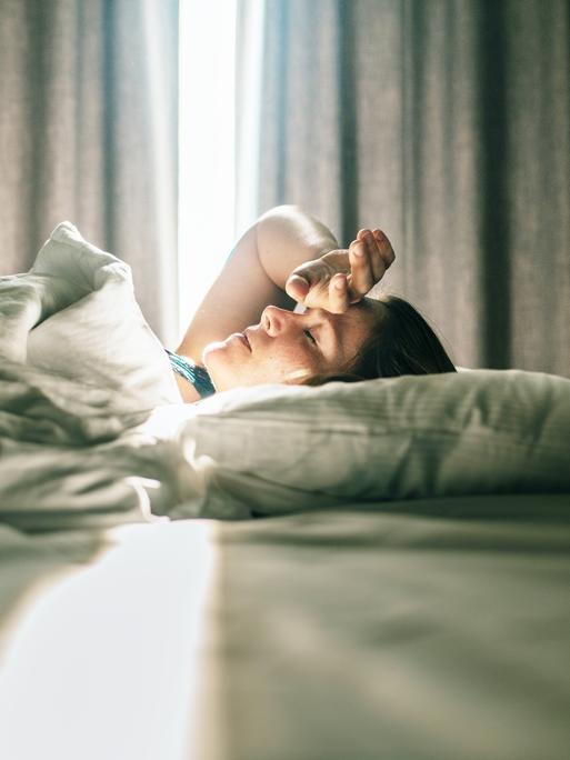 Eine Frau liegt im Bett. Sonniges Tageslicht fällt durch die Vorhänge im Hintergrund. Sie hatte eine Hand an die Stirn gelegt. Eine Frau liegt im Bett. Sonniges Tageslicht fällt durch die Vorhänge im Hintergrund. Sie hatte eine Hand an die Stirn gelegt.