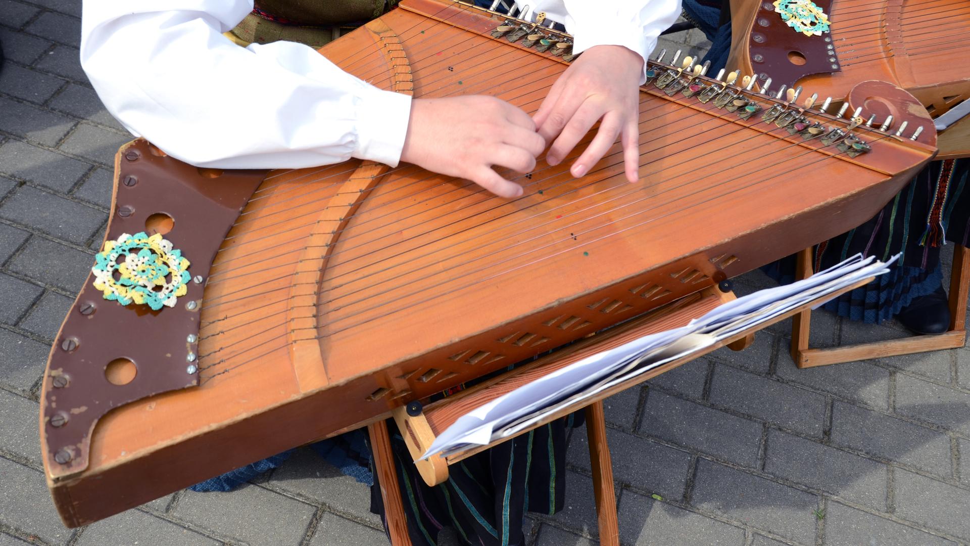 Eine junge Frau in Tracht hat das Zitter-artige Instrumente auf dem Schoß und hat die Hände zum Spiel auf die Saiten gelegt.