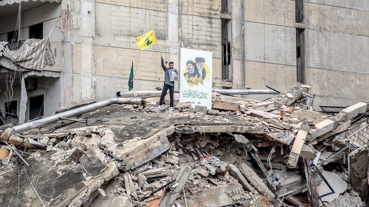 Ein Anhänger der Hisbollah auf einem völlig zerstörten Haus mit wehender Fahne und Porträtfotos. 