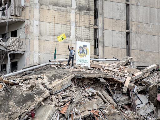 Ein Anhänger der Hisbollah auf einem völlig zerstörten Haus mit wehender Fahne und Porträtfotos. 