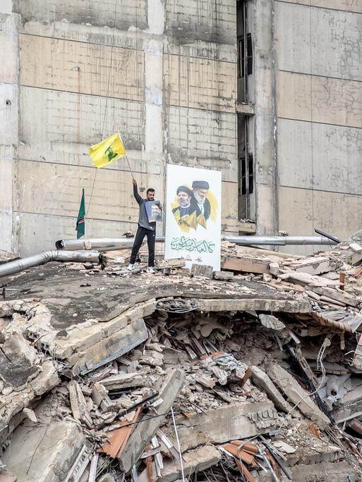 Ein Anhänger der Hisbollah auf einem völlig zerstörten Haus mit wehender Fahne und Porträtfotos. 