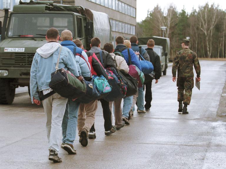 Junge Männer gehen angeführt von einem Soldaten in Tarnanzug an Militärfahrzeugen vorbei.